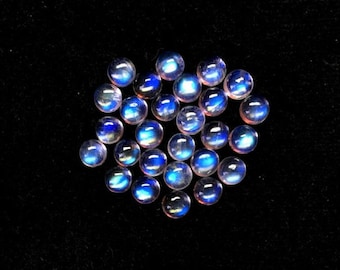 Puede incluir: Un grupo de 18 cabujones de piedra de luna azul iridiscente, cada uno con una superficie lisa y redondeada. Las piedras están dispuestas en un grupo suelto sobre un fondo negro.