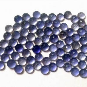 May include: A collection of round, faceted, purple gemstones. The stones are arranged in a loose pile, with some overlapping. The stones are all approximately the same size and shape.