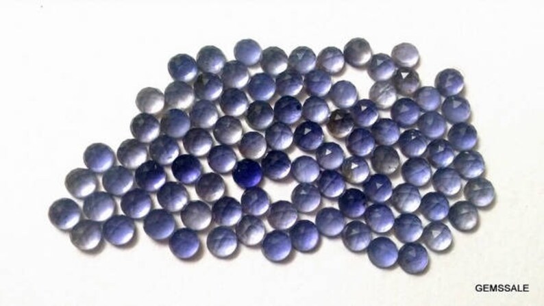 May include: A collection of round, faceted, purple gemstones. The stones are arranged in a loose pile, with some overlapping. The stones are all approximately the same size and shape.