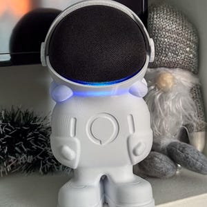 Può includere: Altoparlante bianco a forma di astronauta con una griglia dell'altoparlante nera e una luce blu attorno al bordo del casco.