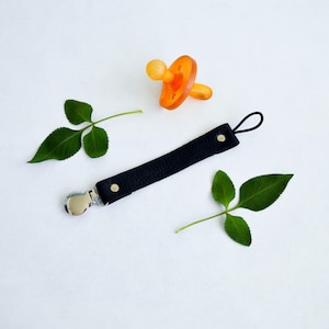 May include: An orange baby pacifier and a black leather pacifier clip with a silver metal clip are displayed on a white surface. Two green leaves are also present. The pacifier clip has gold rivets.