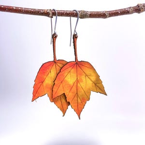 May include: A pair of dangle earrings featuring two orange and yellow leather maple leaves.