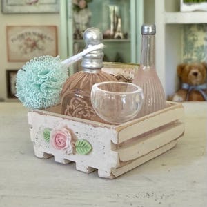 May include: A small white wooden crate with a pink rose and green leaf embellishment containing a light blue bath pouf, a round glass bottle with a silver cap, a clear glass cup, and a tall, slender pink glass bottle with a silver cap.