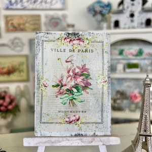 May include: A white wooden sign with a distressed finish featuring a vintage floral illustration and the text "VILLE DE PARIS" and "NOUVEAUTES". The sign is displayed on a white easel.