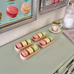 May include: Four small trays of pastel-colored macarons, each tray containing three macarons. The macarons are arranged in a row, with the colors alternating between pink, green, and yellow. The trays are made of a light brown material and are sitting on a light green surface.