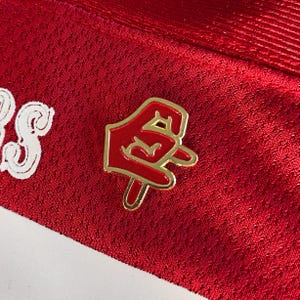 Puede incluir: Un pin de esmalte rojo y dorado que representa un gesto con la mano. El pin está colocado sobre una tela roja texturizada. El diseño del pin incluye una mano roja con un contorno dorado.