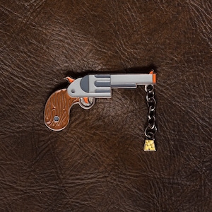 Könnte beinhalten: Eine silberne und braune Emaille-Anstecknadel mit einem Revolver, einer Kette und einem kleinen gelben Anhänger.
