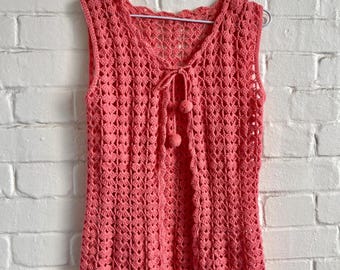 Può includere: Un top all'uncinetto senza maniche color corallo, con scollatura smerlata e scollo a V. Il top presenta una chiusura con fiocco con due piccoli pompon e un delicato motivo traforato. L'indumento è appeso a una gruccia di metallo contro un muro di mattoni bianchi.