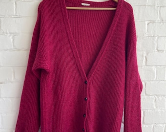 May include: A deep magenta cardigan sweater with a V-neck and long sleeves hangs on a wooden hanger. The sweater has a ribbed knit texture and is buttoned with black buttons. The background is a white brick wall.