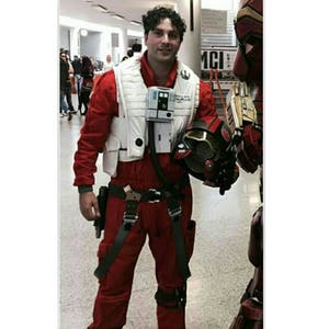 May include: A person wearing a red and white flight suit with a black belt and a helmet. The person is standing in a room with other people.