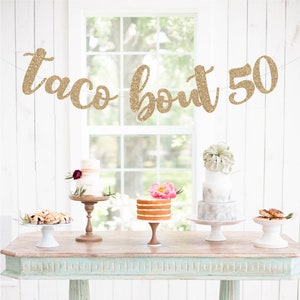 May include: A rustic table displays several small cakes and pastries. A banner above reads "taco bout 50" in gold glitter. The table is painted in a distressed turquoise color, and the background is a white wall and window.
