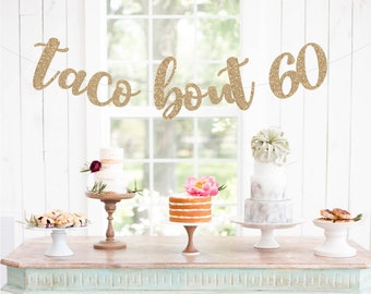 May include: A gold glitter banner with the text "taco bout 60" hanging above a table with four cakes on cake stands. The table is painted a light blue color and has a distressed finish.