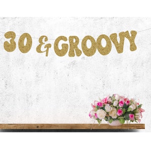 May include: Gold glitter banner with the text "30 & GROOVY" hanging on a white wall. A bouquet of pink and white flowers is on a wooden shelf below the banner.