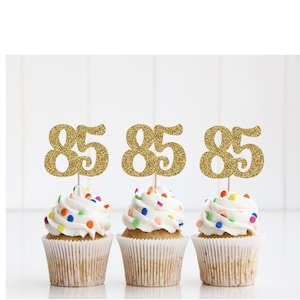 May include: Three cupcakes with white frosting and colorful sprinkles. Each cupcake has a gold glitter "85" cake topper. The cupcakes are in white paper liners.