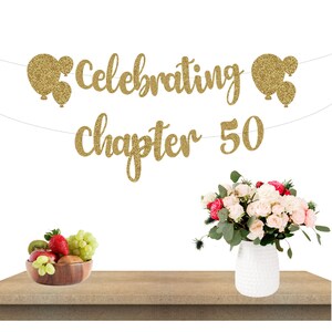 May include: Gold glitter banner with the text "Celebrating chapter 50". The banner is strung with gold glitter balloons on each end. The banner is hanging above a wooden table with a vase of pink and white roses.