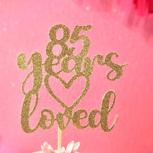 May include: Gold glitter cake topper with the text "85 years loved" and a heart. The topper is on a white cake with pink flowers.