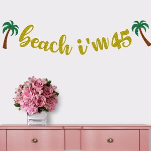May include: Gold glitter banner with the text "beach i'm 45" and two green palm trees on either side.