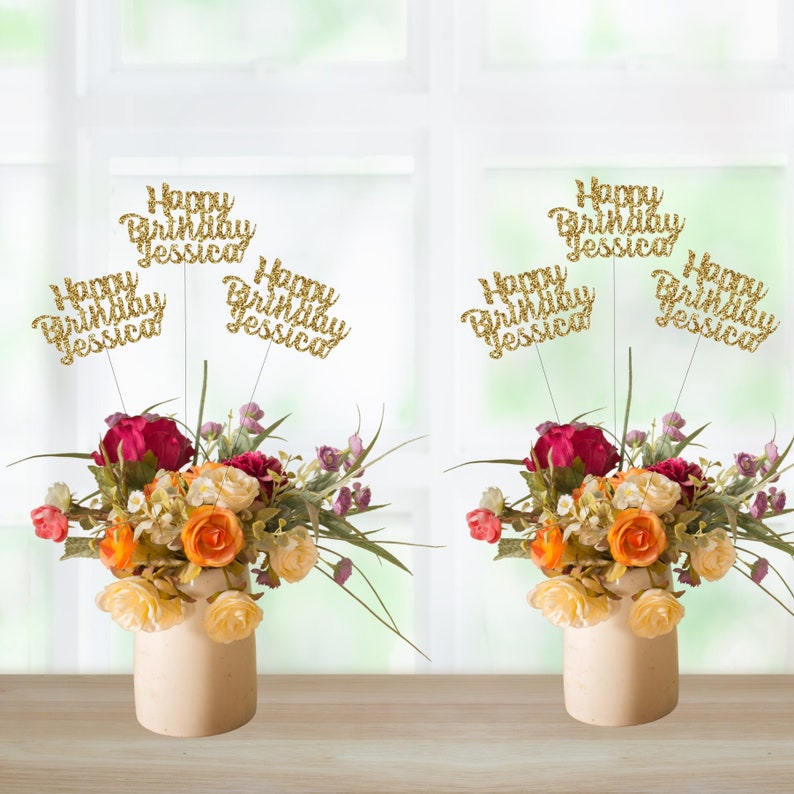 May include: Six gold glitter Happy Birthday Jessica cake toppers with a floral arrangement in two white vases on a wooden table.