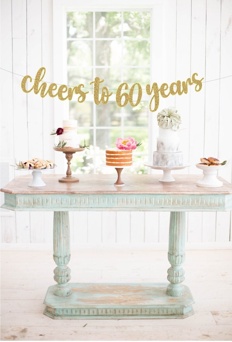 May include: A gold glitter banner with the text "cheers to 60 years" hanging above a table with four cakes on cake stands. The table is painted a light blue color and has a distressed finish.