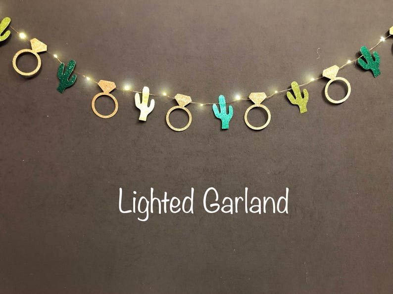 K&ouml;nnte beinhalten: Eine beleuchtete Girlande mit goldenen Diamantring- und Kaktus-Ausschnitten. Die Girlande ist mit kleinen Lichtern versehen, die eine festliche und dekorative Pr&auml;sentation erzeugen. Der Text "Lighted Garland" ist in Wei&szlig; geschrieben.