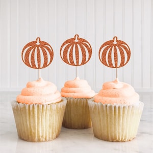 May include: Three cupcakes with orange frosting topped with glitter pumpkin cupcake toppers.