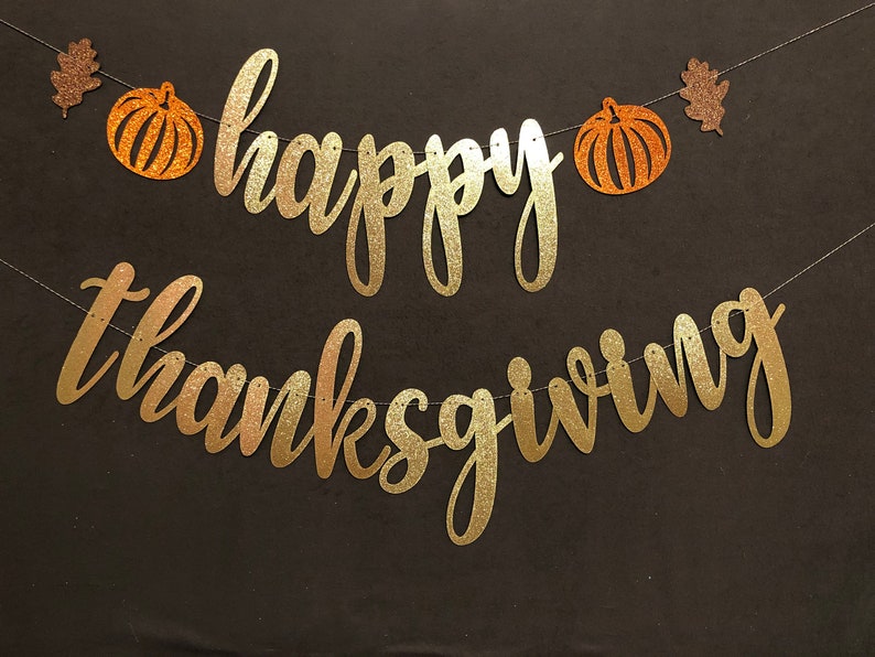 Puede incluir: Una pancarta dorada con purpurina con las palabras "happy thanksgiving" y dos calabazas naranjas con purpurina con hojas marrones con purpurina.