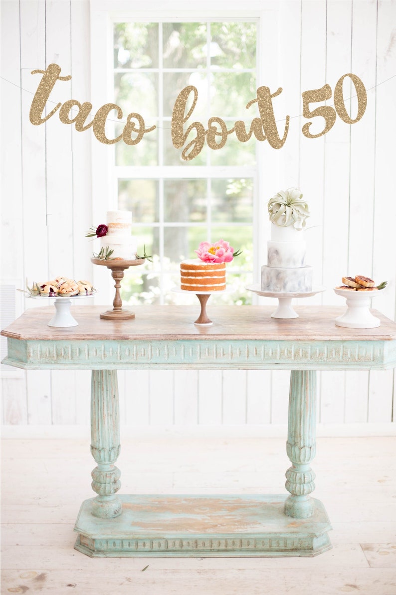 May include: A rustic table displays several small cakes and pastries. A banner above reads "taco bout 50" in gold glitter. The table is painted in a distressed turquoise color, and the background is a white wall and window.
