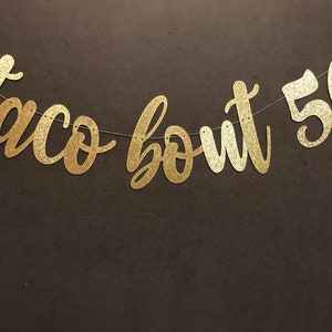 May include: A gold glitter banner that reads "taco bout 50" in a cursive font. The banner is strung on a thin silver string and is set against a dark brown background. Perfect for a 50th birthday celebration.