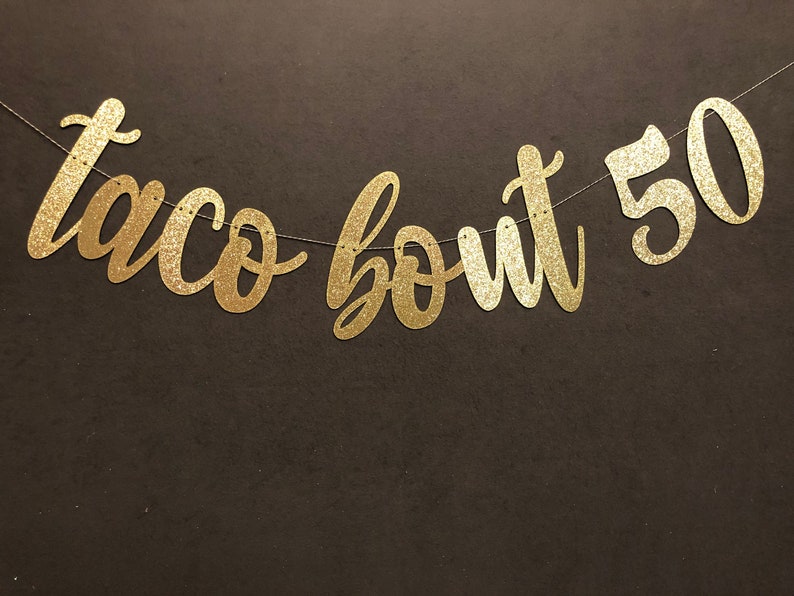 May include: A gold glitter banner that reads "taco bout 50" in a cursive font. The banner is strung on a thin silver string and is set against a dark brown background. Perfect for a 50th birthday celebration.