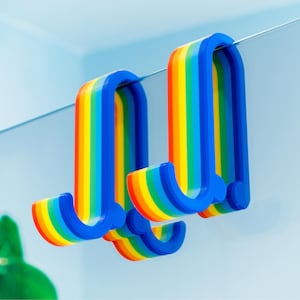 May include: Two rainbow-coloured hooks made of plastic. The hooks are shaped like a letter J and are attached to a wall.