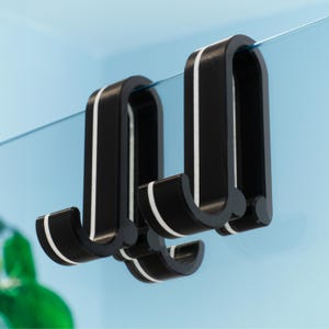 May include: Two black hooks with white stripes, designed to hang over a glass surface. The hooks are curved and have a modern design.