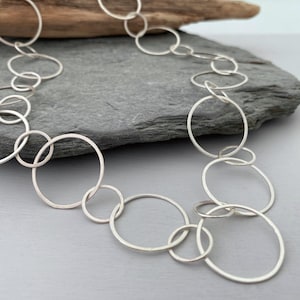 May include: A silver necklace made of interlocking circles of varying sizes. The necklace is displayed on a grey stone surface.