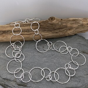 May include: A silver necklace with a design of interconnected hammered circles of different sizes. The necklace is shown on a grey surface with a piece of driftwood in the background. The circles are linked together to form a chain.