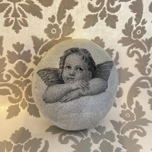 May include: A grey stone featuring a black and white image of a cherub with wings. The cherub's arms are crossed. The stone is set against a background with a floral pattern.