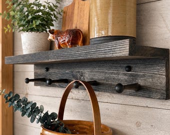May include: A rustic wooden shelf with a dark gray finish, featuring a small potted plant, a decorative cow figurine, and a wooden cutting board. Below, a row of black hooks holds a woven basket with a handle. The shelf is mounted on a light wood-paneled wall.