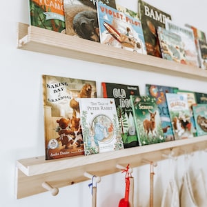 Pu&ograve; includere: Uno scaffale di legno con ganci sotto, che contiene una collezione di libri per bambini. I libri hanno copertine colorate con varie illustrazioni, tra cui animali, un aereo e una scena di cottura. Il testo sui libri include titoli come "Shh! Bears Sleeping", "Violet Pilot", "Baking Day at Grandma's", e "The Tale of Peter Rabbit".