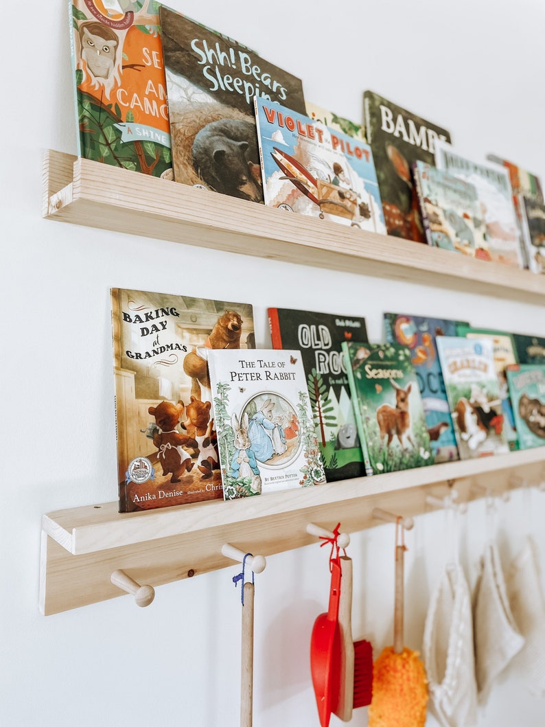 Pu&ograve; includere: Uno scaffale di legno con ganci sotto, che contiene una collezione di libri per bambini. I libri hanno copertine colorate con varie illustrazioni, tra cui animali, un aereo e una scena di cottura. Il testo sui libri include titoli come "Shh! Bears Sleeping", "Violet Pilot", "Baking Day at Grandma's", e "The Tale of Peter Rabbit".
