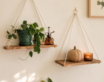 May include: Two hanging wooden shelves with plants and decor. One shelf holds a green potted plant and a brown glass mister. The other shelf displays a woven pumpkin and a lit candle in a glass holder. The shelves are suspended by white rope.