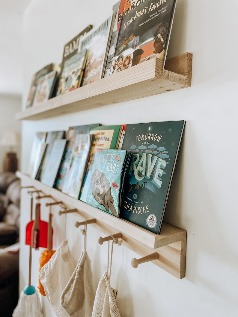 Pu&ograve; includere: Uno scaffale in legno con ganci sotto, che contiene una collezione di libri per bambini. I libri sono disposti su due file, la fila superiore presenta titoli come "Grandma's Favorite" e "The Big Boy", e la fila inferiore presenta titoli come "I Love You Baby" e "Tomorrow I'll Be Rave".