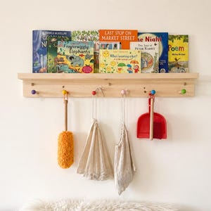 May include: A wooden shelf with colorful books and a row of pegs. Below, a yellow duster, two beige cloths, and a red dustpan hang. The books include titles like "Fringerwiggly Elephants" and "Last Stop on Market Street".