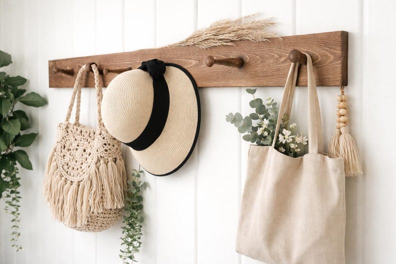 May include: A wooden wall-mounted coat rack with four pegs, holding a beige macrame bag, a straw hat with a black band, and a beige tote bag filled with greenery. A decorative tassel hangs on the right.