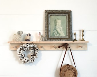 May include: A wooden shelf with hooks, a cotton wreath, a small woven bag, and a framed photo of a young girl. The shelf is decorated with small figurines of sheep and a llama.