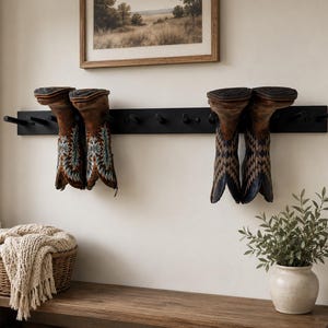 May include: A long, horizontal black wall-mounted rack featuring multiple cylindrical pegs. Two pairs of brown leather cowboy boots with intricate stitching are hanging upside down on the pegs, with several empty pegs remaining on the rack.
