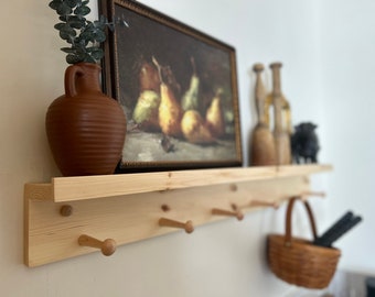 May include: A wooden shelf with five hooks, a brown clay vase with green leaves, a framed painting of pears, two brown bottles, and a black metal object. The shelf is mounted on a white wall.