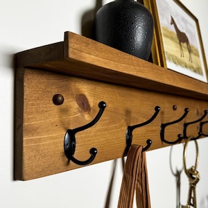 May include: A wooden shelf with five black metal hooks. The shelf is stained a dark brown colour and has a decorative top edge. A brown leather strap hangs from one of the hooks.