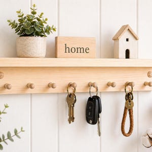 Könnte beinhalten: Ein heller Holzschlüsselhalter mit einem Regal, Haken und dem Wort "home" auf einem Block. Ein kleines Haus und eine Topfpflanze stehen auf dem Regal. Schlüssel hängen an den Haken. Der Schlüsselhalter ist an einer weißen Wand befestigt.