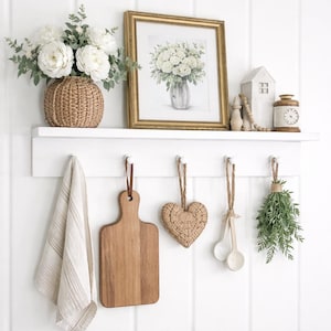 May include: A white wall shelf with decorative items. A framed floral print, a woven vase with white flowers, and a small house figurine are on the shelf. Below, a wooden cutting board, a heart-shaped woven ornament, and a bundle of herbs hang from hooks.