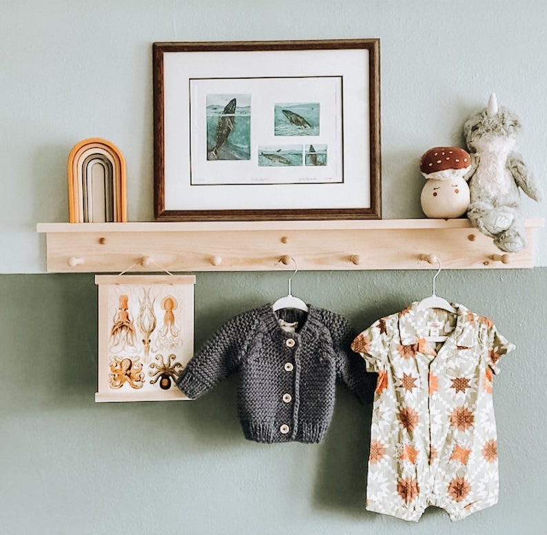 K&ouml;nnte beinhalten: Ein Holzregal mit Haken, ein gerahmtes Bild von Walen, ein regenbogenfarbener Holzbogen, ein ausgestopftes Narwal, ein pilzf&ouml;rmiges Spielzeug und zwei Kinderoutfits, die an den Haken h&auml;ngen. Ein Outfit ist ein dunkelgrauer Cardigan und das andere ist ein wei&szlig;er und brauner Blumenprint-Strampler.