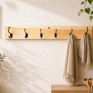 May include: A wooden wall-mounted coat rack with six black hooks, holding two beige fringed throws. A framed sign with the text "SIMPLE BEAUTIFUL EVERYDAY home" hangs on the wall. Decorative plants in woven baskets and pots complete the scene.