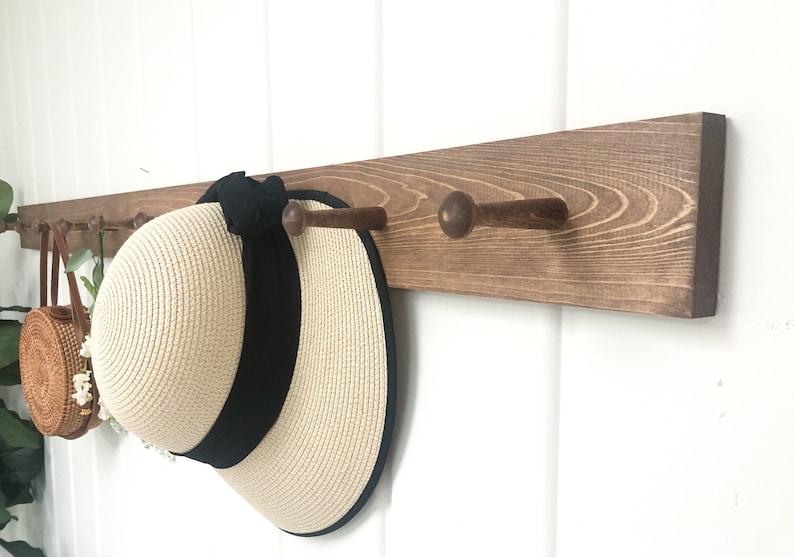 May include: A wooden wall-mounted coat rack with three dark brown pegs. A beige sun hat with a black band hangs from one peg. A small round woven bag with a leather strap hangs from the left side.
